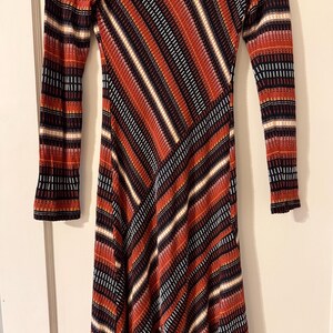 Puede incluir: Un vestido de manga larga con un llamativo estampado de rayas diagonales en tonos rojos, naranjas, negros y blancos. El vestido tiene una silueta ajustada y un tejido de punto texturizado.