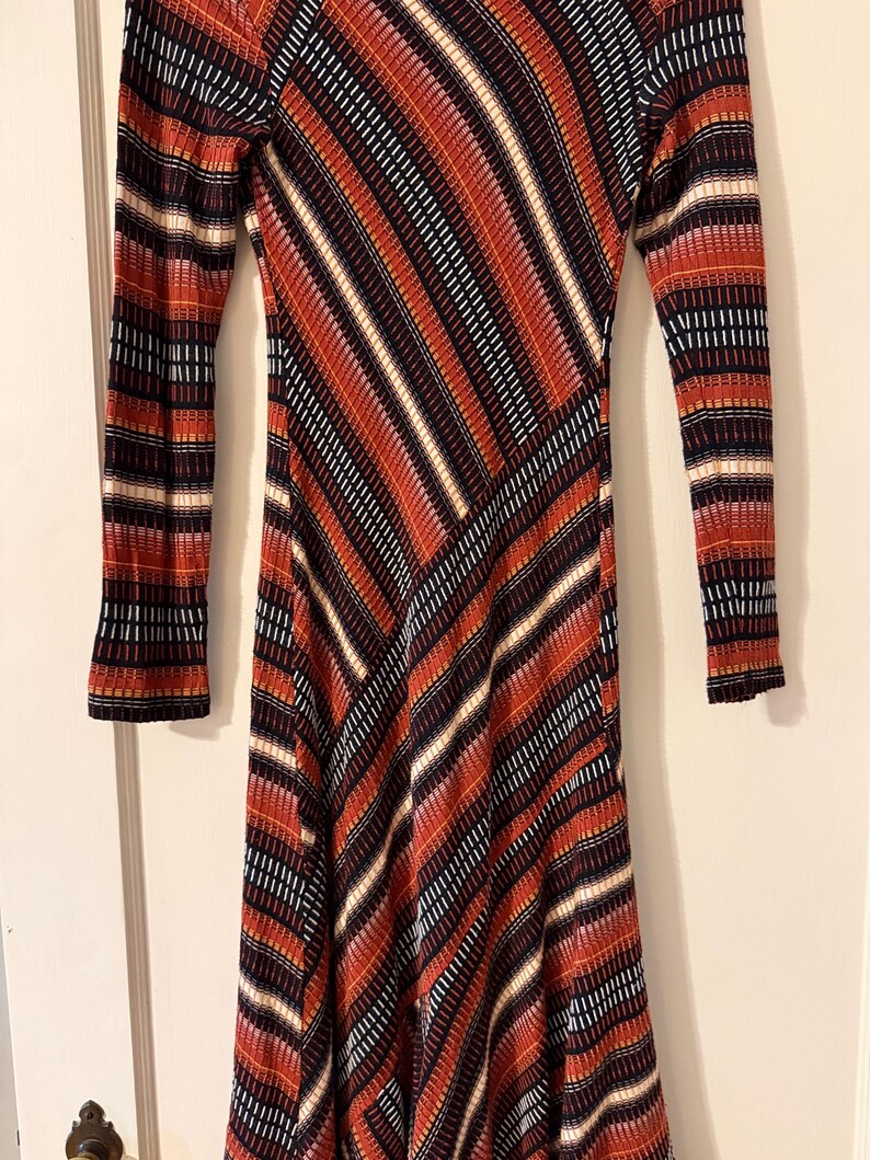 Puede incluir: Un vestido de manga larga con un llamativo estampado de rayas diagonales en tonos rojos, naranjas, negros y blancos. El vestido tiene una silueta ajustada y un tejido de punto texturizado.