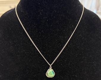 May include: A silver necklace with a triangular pendant. The pendant has a green and blue stone set in a silver frame. The chain is delicate and hangs on a black background.