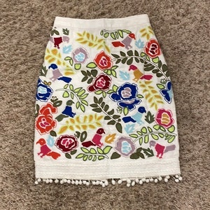 May include: White skirt with a colorful floral and bird embroidery pattern. The skirt has a white pom-pom trim.