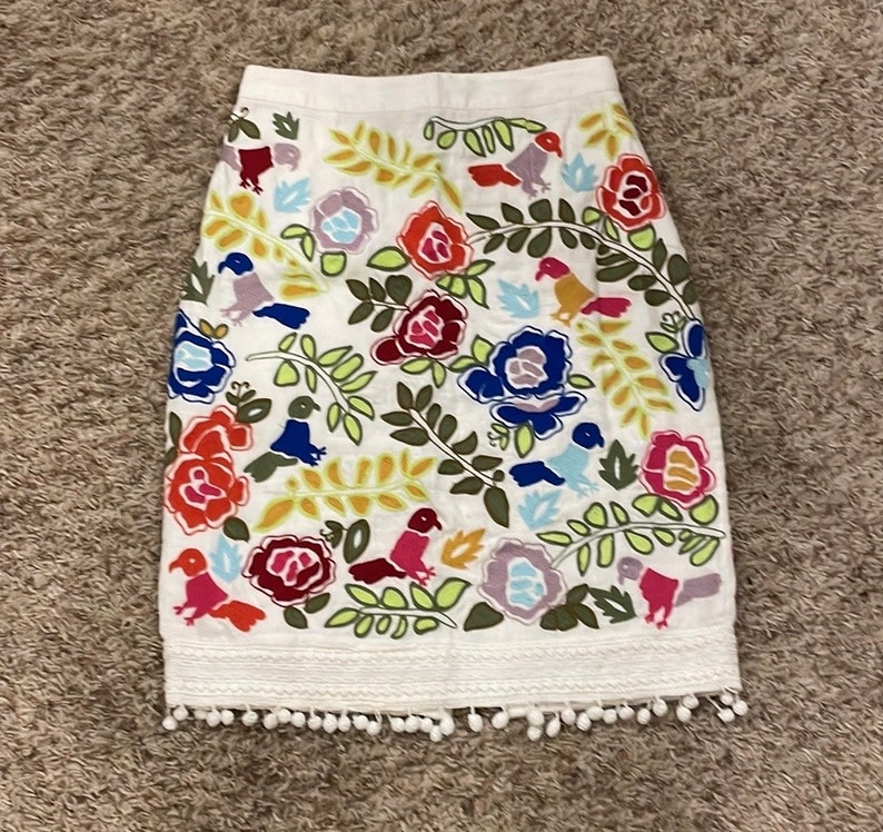 May include: White skirt with a colorful floral and bird embroidery pattern. The skirt has a white pom-pom trim.