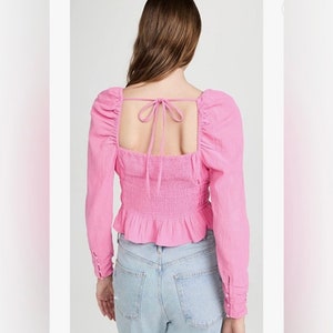 May include: A pink long-sleeved blouse with a square neckline and a tie at the back. The blouse has a fitted bodice and a ruffled hem.