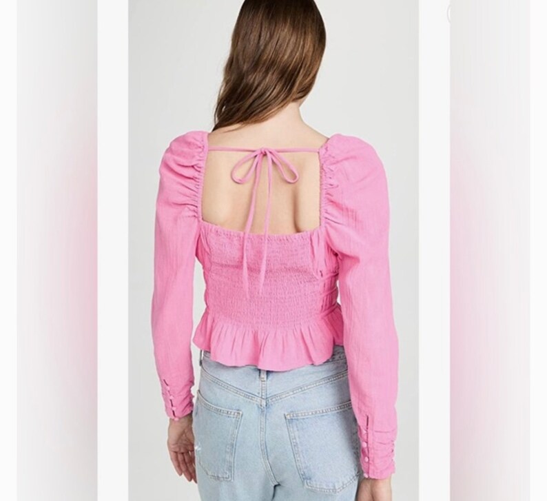 May include: A pink long-sleeved blouse with a square neckline and a tie at the back. The blouse has a fitted bodice and a ruffled hem.