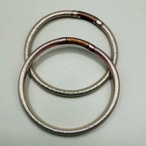 May include: Two silver-colored bangles with a textured surface are displayed. Each bangle has a copper-colored section with the word "BLESSED" engraved on it. The bangles are circular and appear to be made of metal.