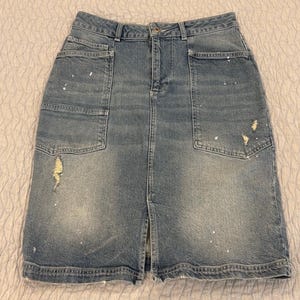 May include: A light blue denim skirt with a front slit and two large front pockets. The skirt has distressed details and white paint splatters. The skirt is a knee-length style.