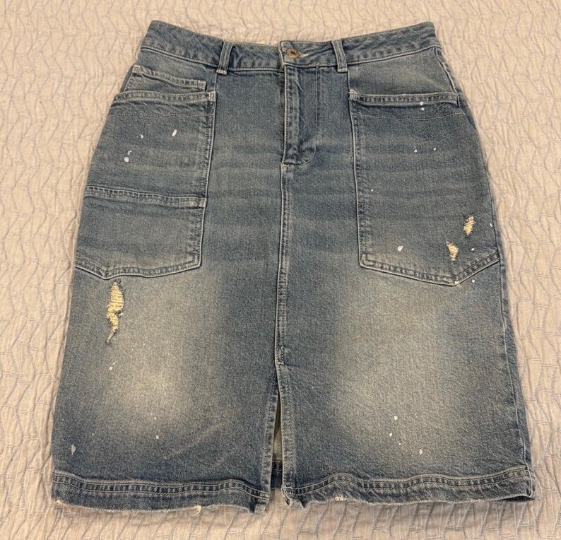 May include: A light blue denim skirt with a front slit and two large front pockets. The skirt has distressed details and white paint splatters. The skirt is a knee-length style.