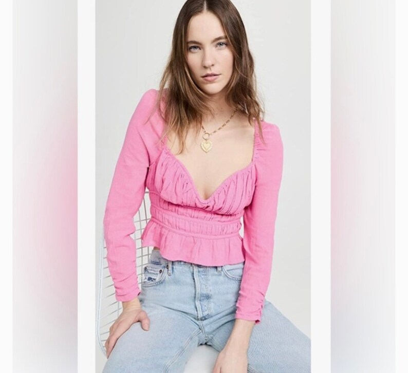 May include: A pink long-sleeved blouse with a gathered neckline and a fitted bodice. The blouse has a ruffled hem and is cinched at the waist.