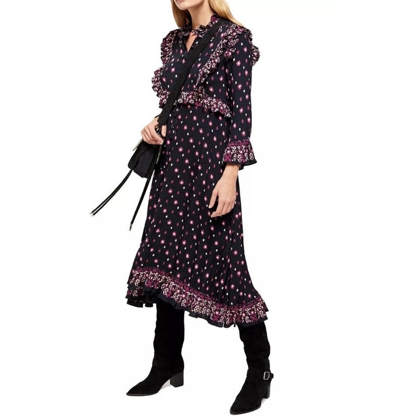 May include: A black midi dress with a pink floral print and ruffle detailing. The dress has a long sleeve and a high neck. The model is wearing black boots.