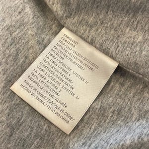 Op de afbeelding: Een wit label met zwarte tekst die de stofsamenstelling van een kledingstuk beschrijft. Het label luidt: RN#104537 CA#32054 SELF/SELF/SELBST/AUTO/AUTO 100% POLYESTER/POLYESTER/POLVESTER/POLIESTERE/POLI&Eacute;STER LINING 1/DOUBLURE 1/FUTTER 1/ LA LINEA 1/FORRO 1 100%RAYON/RAYONNE/KUNSTSEIDE/RAION/RAYON LINING 2/DOUBLURE 2/FUTTER 2/ LA LINEA 2/FORRO 2 100% COTTON/COTON/BAUMWOLLE/COTONE/ALGOD&Oacute;N MADE IN CHINA/Fabriqu&eacute; en Chine/HECHO EN CHINA/FEITO EM CHINA