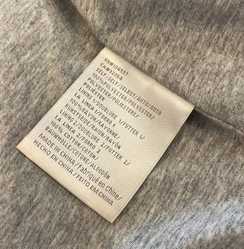 Op de afbeelding: Een wit label met zwarte tekst die de stofsamenstelling van een kledingstuk beschrijft. Het label luidt: RN#104537 CA#32054 SELF/SELF/SELBST/AUTO/AUTO 100% POLYESTER/POLYESTER/POLVESTER/POLIESTERE/POLI&Eacute;STER LINING 1/DOUBLURE 1/FUTTER 1/ LA LINEA 1/FORRO 1 100%RAYON/RAYONNE/KUNSTSEIDE/RAION/RAYON LINING 2/DOUBLURE 2/FUTTER 2/ LA LINEA 2/FORRO 2 100% COTTON/COTON/BAUMWOLLE/COTONE/ALGOD&Oacute;N MADE IN CHINA/Fabriqu&eacute; en Chine/HECHO EN CHINA/FEITO EM CHINA