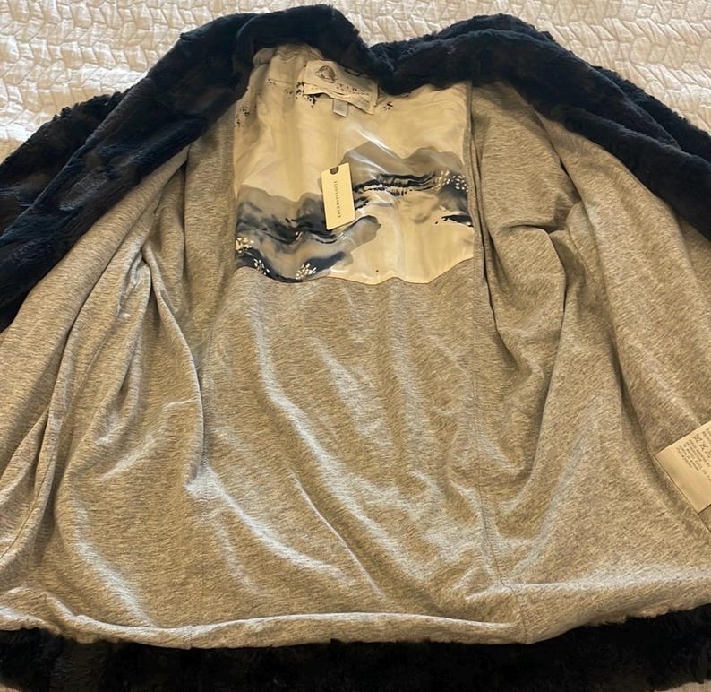 Op de afbeelding: Een zwart nep bontjasje met een grijze voering en een witte en blauwe geprinte stof die de binnenkant van de kraag bekleedt. Het jasje heeft een label met de tekst "Anthropologie" erop.