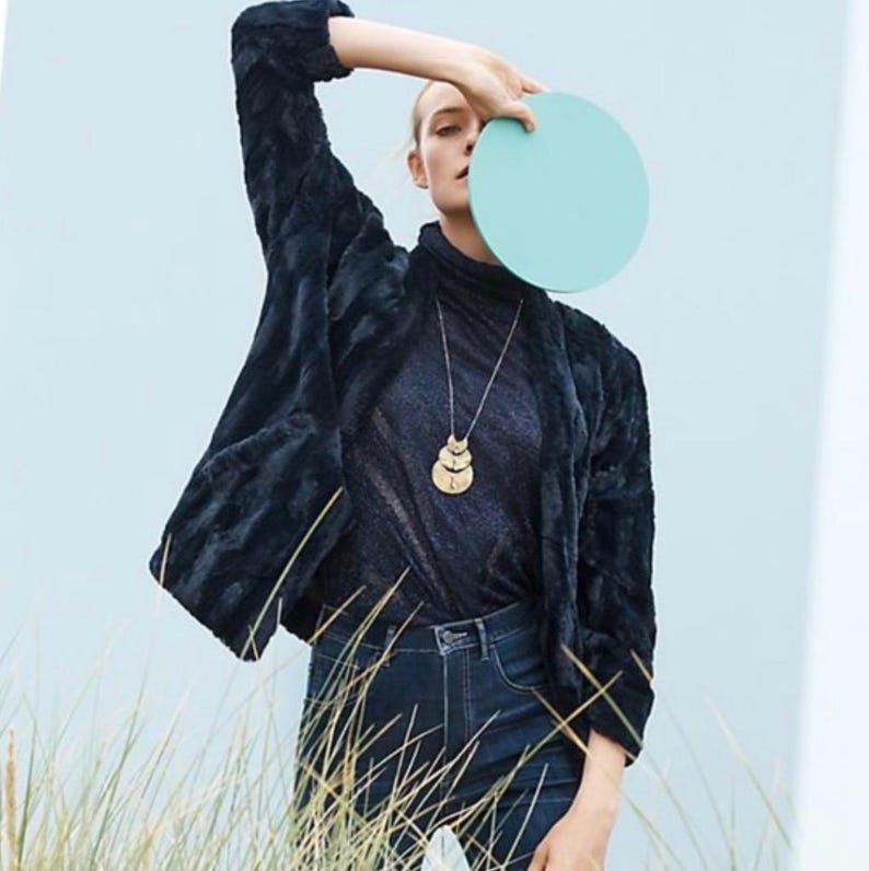 Op de afbeelding: Een vrouw draagt een zwart nep bontjasje, een zwart shirt met een hoge hals, blauwe jeans en een gouden gelaagde ketting. Ze houdt een lichtblauwe cirkel voor haar gezicht.