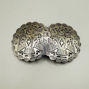 May include: Two silver-toned, scalloped-edge conchos. Each concho features an engraved design of a bird with outstretched wings, surrounded by geometric patterns. The conchos are likely for a belt or other accessory.