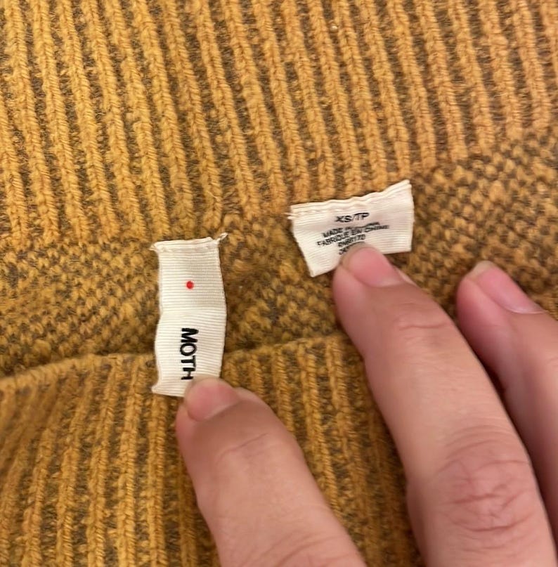 May include: A mustard yellow sweater with a ribbed texture and a small white tag that says "MOTH" on it. The tag is attached to the sweater with a small red dot.