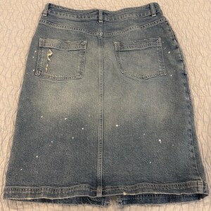 May include: A light blue denim skirt with two back pockets and a distressed detail on one pocket. The skirt has a split at the bottom and white paint splatters throughout.