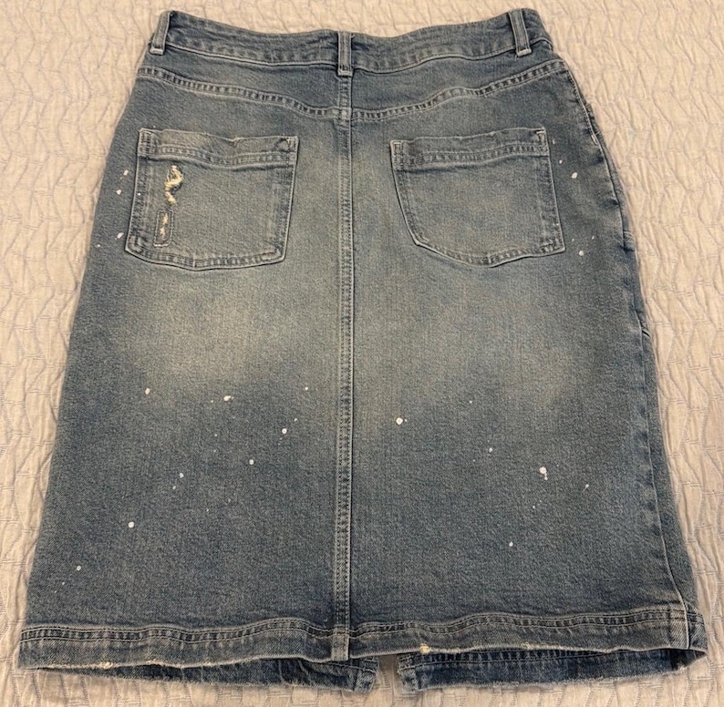 May include: A light blue denim skirt with two back pockets and a distressed detail on one pocket. The skirt has a split at the bottom and white paint splatters throughout.