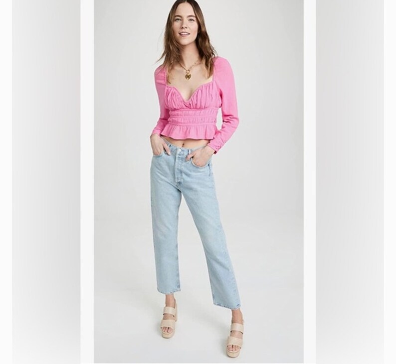 May include: A pink, long-sleeved, cropped blouse with a sweetheart neckline and a gathered waist. The blouse is worn with light blue, straight-leg jeans and beige platform sandals.