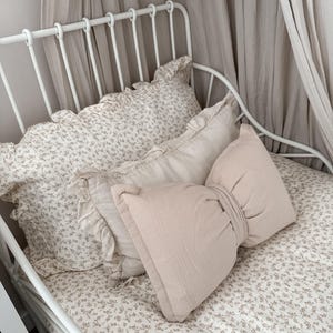 May include: A white metal bed frame with a floral patterned bedspread and pillows. The pillows include a ruffled pillow, a rectangular pillow, and a large bow-shaped pillow in a light pink color. A sheer curtain hangs above the bed.