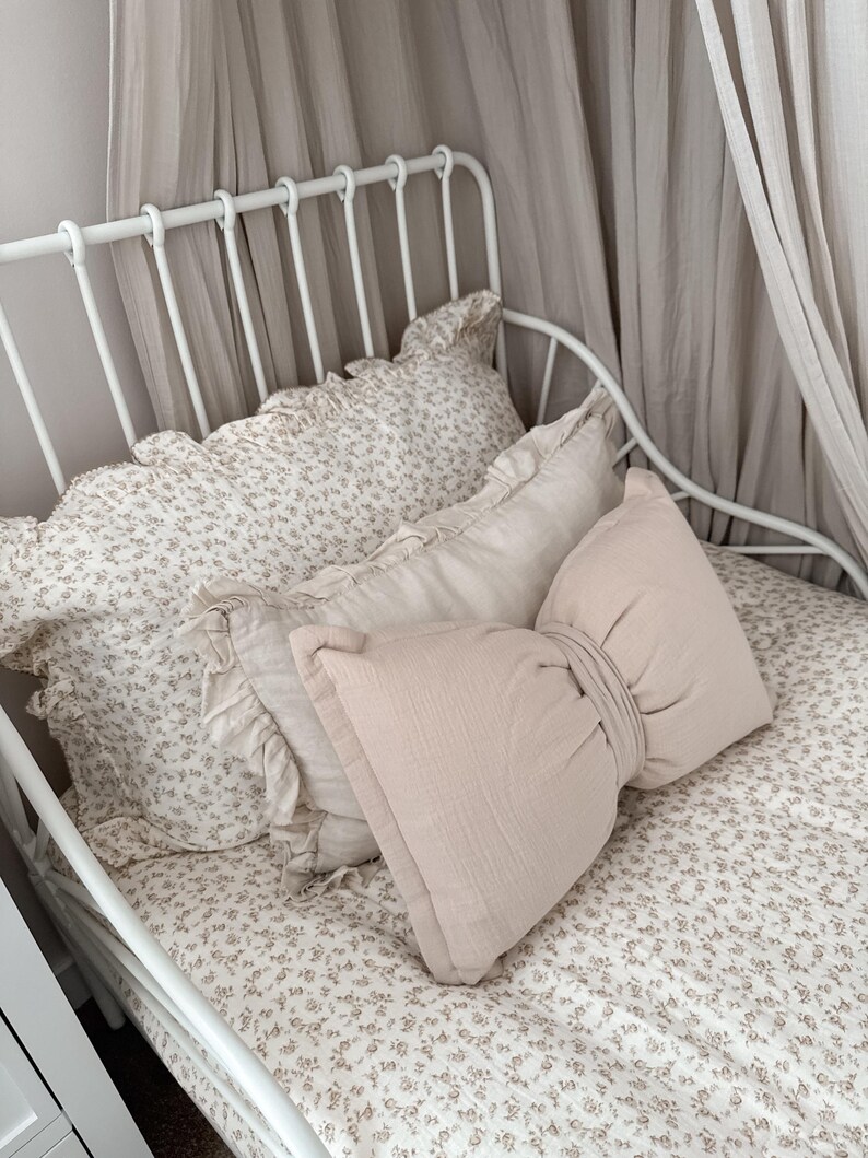 May include: A white metal bed frame with a floral patterned bedspread and pillows. The pillows include a ruffled pillow, a rectangular pillow, and a large bow-shaped pillow in a light pink color. A sheer curtain hangs above the bed.