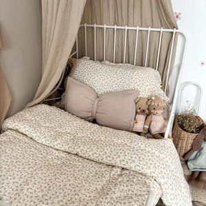 neutral bow cushion sits on a white metal framed bed in this toddlers vintage themed bedroom. The beige bow pillow has been styled with a beige canopy hanging behind the bed and vintage floral bed covers