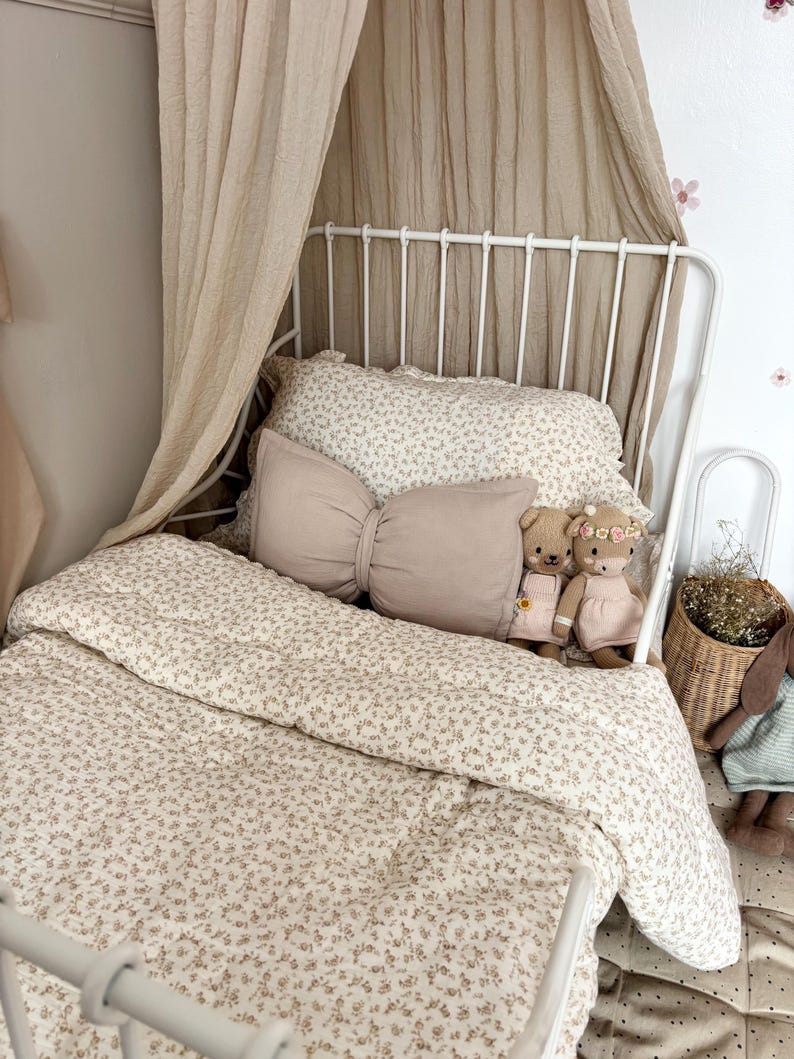 neutral bow cushion sits on a white metal framed bed in this toddlers vintage themed bedroom. The beige bow pillow has been styled with a beige canopy hanging behind the bed and vintage floral bed covers