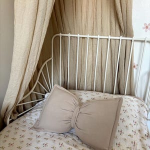 May include: A beige bow-shaped pillow rests on a bed with a floral patterned comforter. The bed has a white metal frame and is draped with a beige canopy. The room has a soft, neutral color palette.
