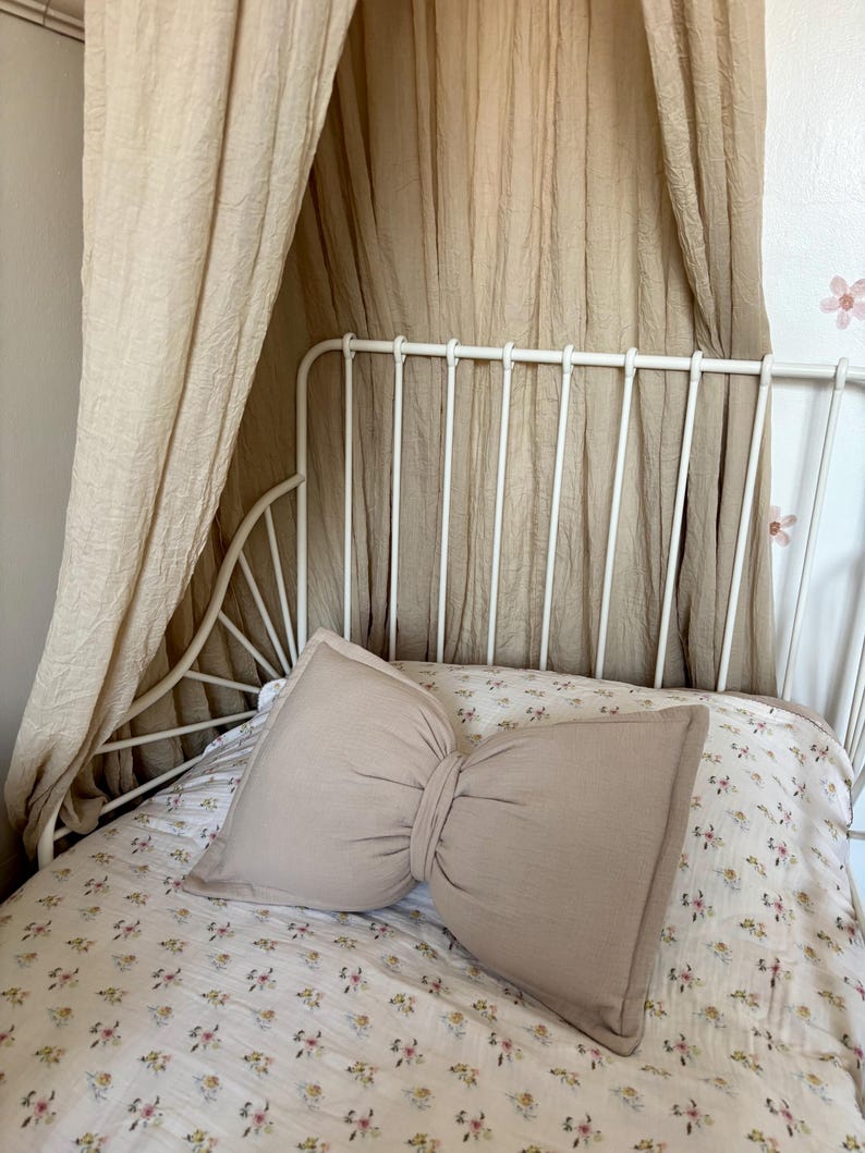 May include: A beige bow-shaped pillow rests on a bed with a floral patterned comforter. The bed has a white metal frame and is draped with a beige canopy. The room has a soft, neutral color palette.