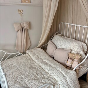 May include: A white metal bed frame with a floral patterned comforter and pillows. A large beige bow hangs on the wall above the bed. Two teddy bears sit on the pillows. A beige canopy drapes over the bed.