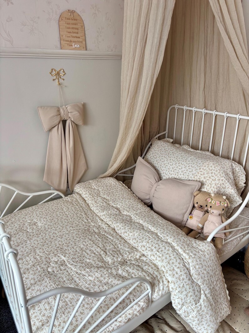 May include: A white metal bed frame with a floral patterned comforter and pillows. A large beige bow hangs on the wall above the bed. Two teddy bears sit on the pillows. A beige canopy drapes over the bed.
