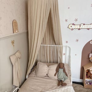 May include: A beige canopy bed with a white metal frame. The bed is made with a beige comforter, a bow-shaped pillow, and a stuffed bunny. The wall is decorated with floral wallpaper and a wooden sign.