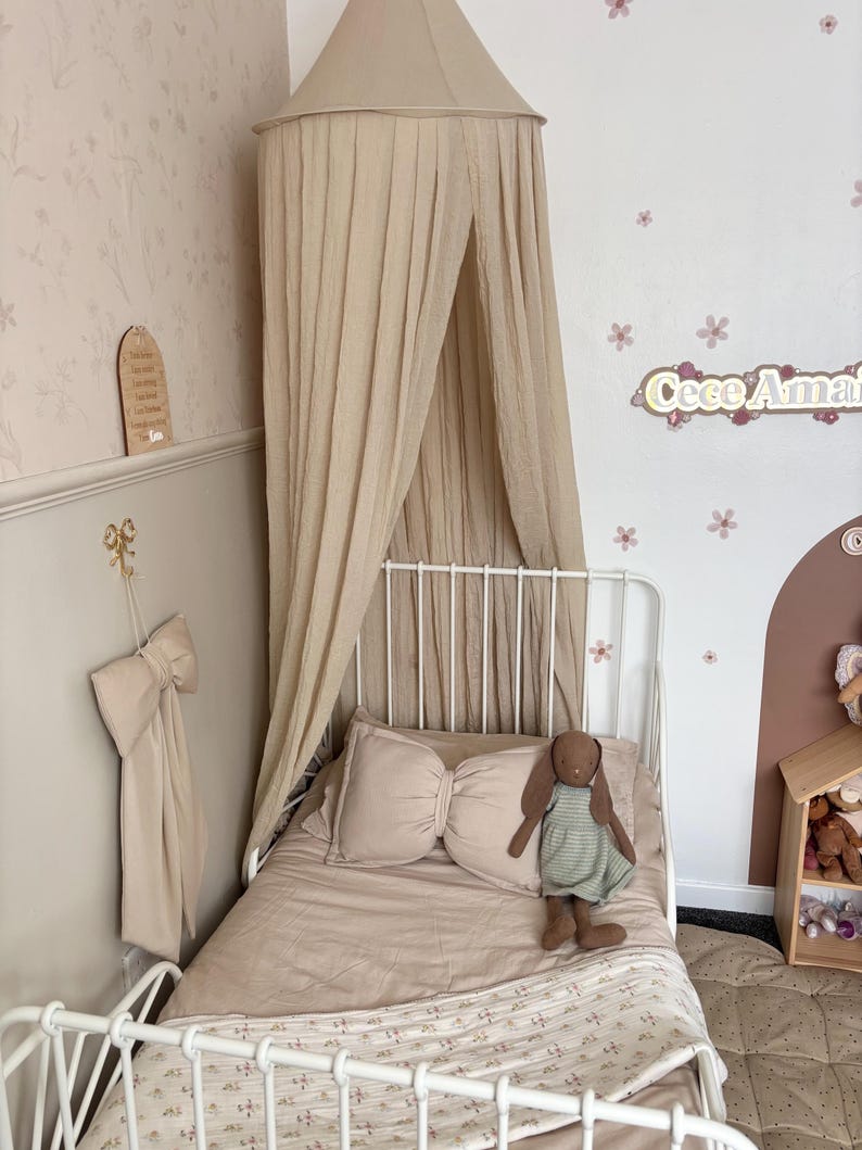 May include: A beige canopy bed with a white metal frame. The bed is made with a beige comforter, a bow-shaped pillow, and a stuffed bunny. The wall is decorated with floral wallpaper and a wooden sign.