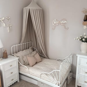 May include: A white metal toddler bed with a floral patterned comforter and pillows. The room features a canopy, two white nightstands, and a wall decoration that says "Olivia". The room is decorated in neutral colors.
