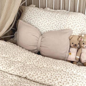 neutral bow cushion sits on a white metal framed bed in this toddlers vintage themed bedroom. The beige bow pillow has been styled with a beige canopy hanging behind the bed and vintage floral bed covers