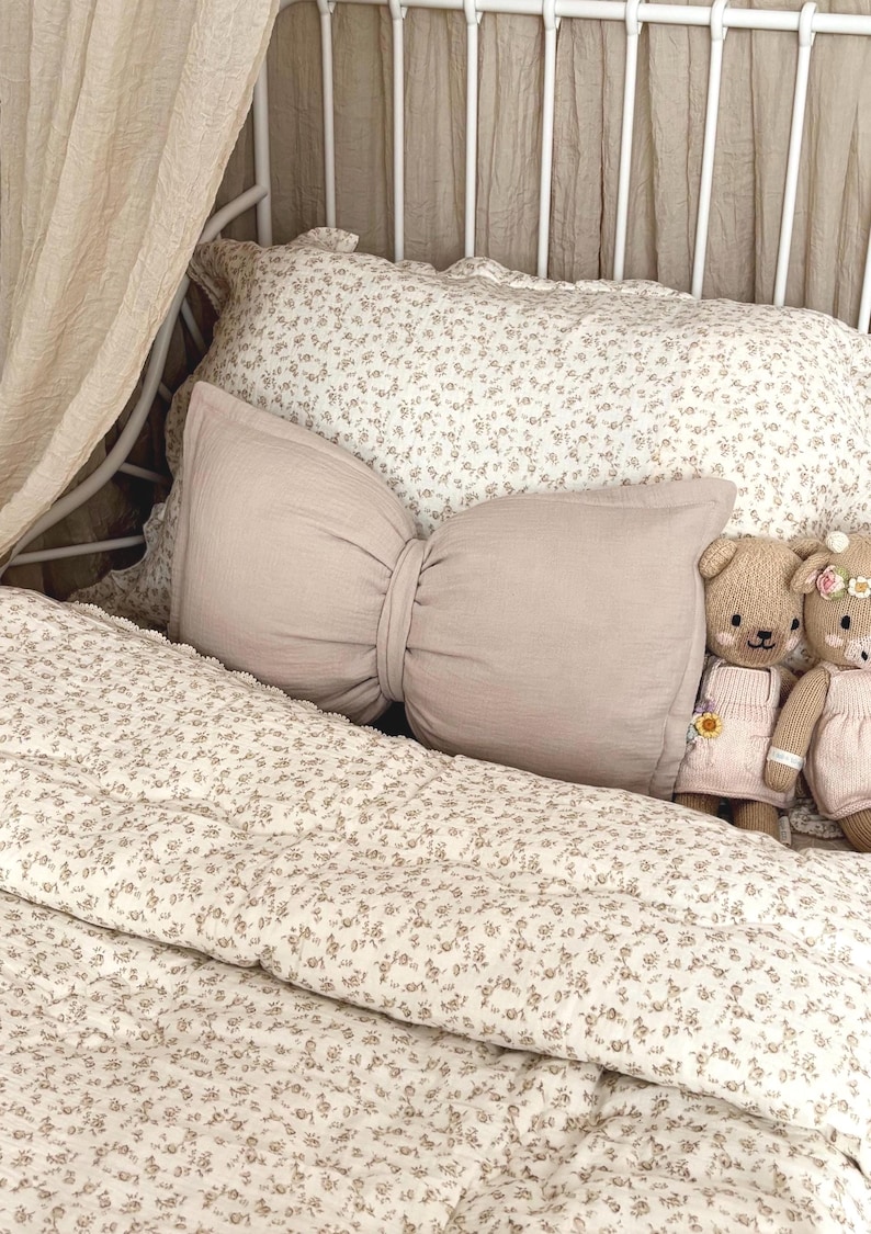 neutral bow cushion sits on a white metal framed bed in this toddlers vintage themed bedroom. The beige bow pillow has been styled with a beige canopy hanging behind the bed and vintage floral bed covers
