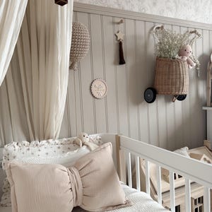 May include: A nursery scene with a white crib, a large bow-shaped pillow, and a decorative wall. The wall features a woven basket with flowers and a stuffed animal, a wooden star, and a hot air balloon mobile. The color palette is soft, with beige and white tones.