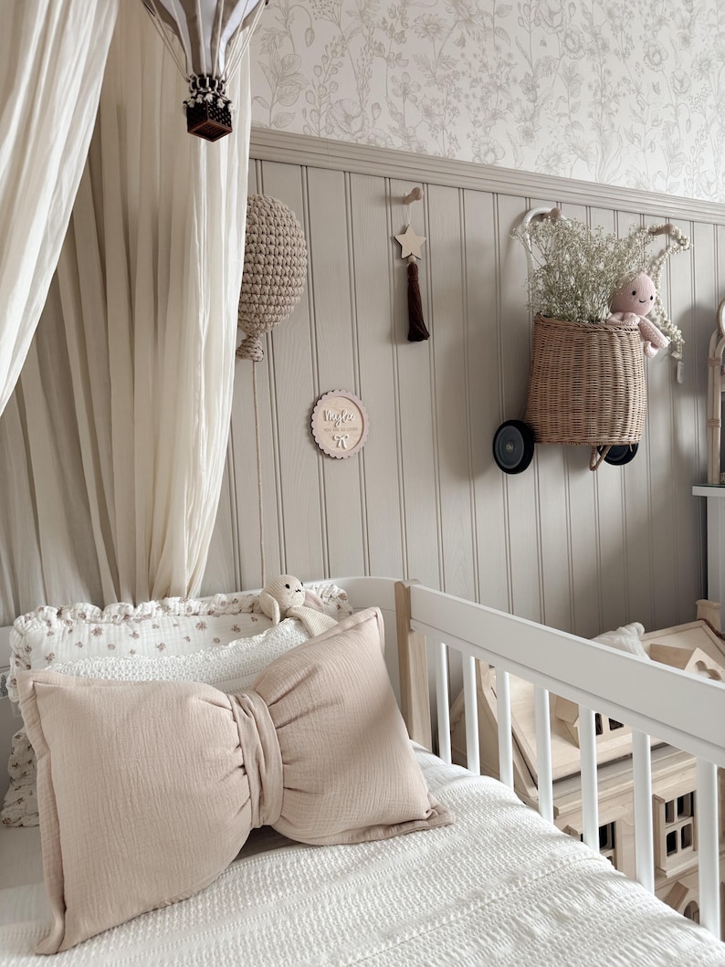 May include: A nursery scene with a white crib, a large bow-shaped pillow, and a decorative wall. The wall features a woven basket with flowers and a stuffed animal, a wooden star, and a hot air balloon mobile. The color palette is soft, with beige and white tones.
