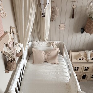 May include: A nursery scene with a white crib, soft bedding, and decorative elements. A beige bow-shaped pillow sits on the bed. A hot air balloon mobile and other wall decorations add to the room's cozy aesthetic.