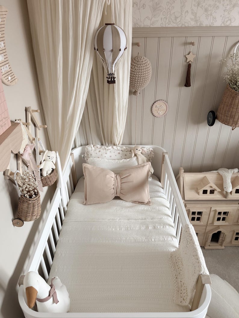 May include: A nursery scene with a white crib, soft bedding, and decorative elements. A beige bow-shaped pillow sits on the bed. A hot air balloon mobile and other wall decorations add to the room's cozy aesthetic.