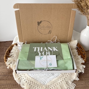 May include: A brown cardboard box with the logo "Sentimentally Handmade" is open, revealing a gift box with green tissue paper and a "Thank You" card. The box is in a woven basket with a white fringed cloth.