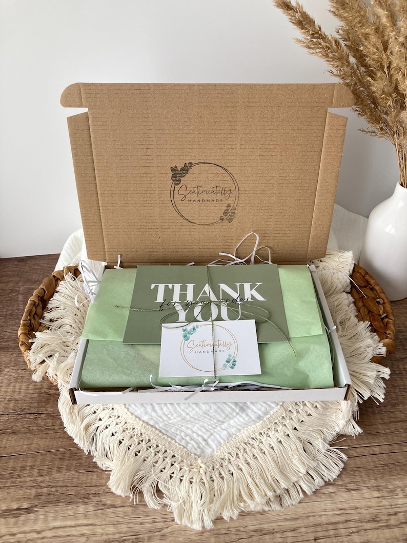 May include: A brown cardboard box with the logo "Sentimentally Handmade" is open, revealing a gift box with green tissue paper and a "Thank You" card. The box is in a woven basket with a white fringed cloth.