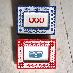 May include: Two small rectangular picture frames with hand-painted designs. The top frame is white with blue floral patterns and contains a drawing of three red horseshoes. The bottom frame is white with red floral patterns and a blue camera.