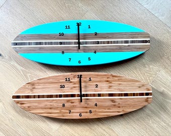 May include: Two surfboard-shaped wall clocks with black hands and numerals. The top clock features bright turquoise ends with a central wood grain and white striped pattern. The bottom clock is made of natural bamboo with a central dark wood stripe.