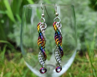 May include: Rainbow coloured metal chain earrings with silver hooks. The earrings are hanging from a wine glass.
