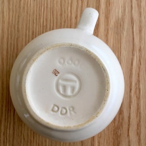 Pu&ograve; includere: Tazza in ceramica bianca con manico. Il fondo della tazza ha un timbro marrone con il testo "DDR" e un simbolo con il numero "963" e "32".