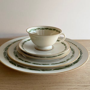 Puede incluir: Un juego de cuatro platos de cerámica blanca con un diseño de hojas verdes y doradas. Los platos están apilados uno encima del otro, con una pequeña taza de té blanca con un diseño de hojas verdes y doradas en la parte superior.