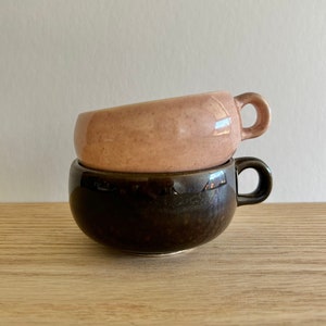 May include: Two ceramic mugs stacked vertically. The top mug is a speckled light pink with a glossy finish and a small round handle. The bottom mug is a solid dark brown with a smooth, reflective surface and a matching round handle.