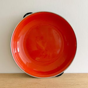May include: A vibrant orange enamel bowl with a silver rim and black handles. The bowl is round and sits on a wooden surface. The interior of the bowl is a bright, glossy orange.