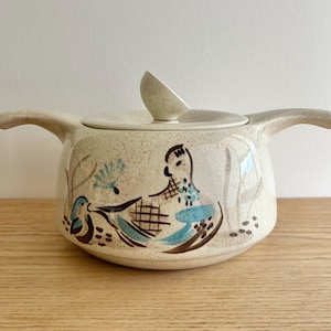 Peut inclure: Plat à casserole en céramique crème avec couvercle et deux poignées. Le plat présente un motif peint à la main d'oiseaux dans des tons de bleu et de marron. Le couvercle a une petite poignée en forme de cuillère.