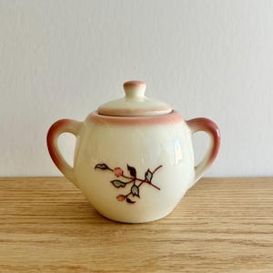 May include: A small, cream-colored ceramic sugar bowl with a lid and two handles. The rim and handles are a reddish-pink color. A floral design with pink and green accents decorates the front. The sugar bowl is sitting on a wooden surface.