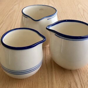 Pu&ograve; includere: Tre brocche di crema in ceramica bianca con bordo blu. Le brocche sono di dimensioni diverse e hanno una forma arrotondata.